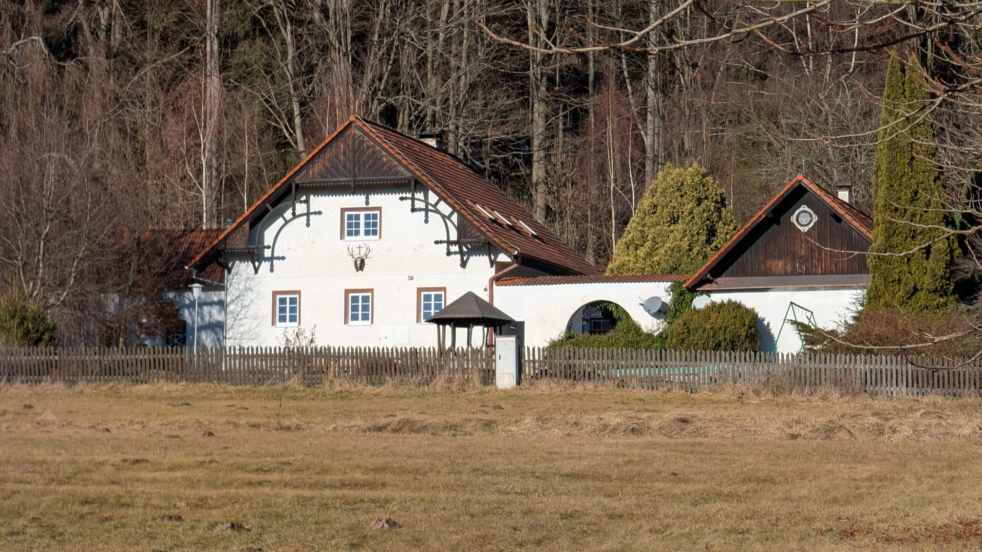 Myslivna Pod Kohoutem – bílé stavení s dřevěnými štíty na okraji lesa